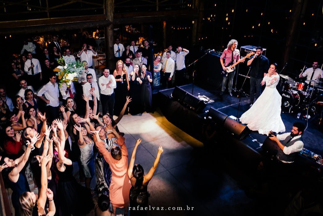 Noiva jogou o buquê e está olhando de cima do palco pra ver quem vai pegar enquanto que no chão várias meninas tentam pegar o buquê.